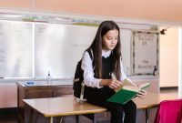 Young girl sitting on desk in classroom reading a book, wearing a backpack.