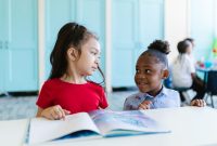 Two young children in a classroom interact while reading a book together.