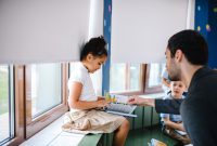A teacher helps a young girl read a book with fellow students in a classroom setting.