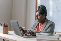 African American female teacher in formal outfit sitting at table in classroom and typing on laptop and taking notes on notepad