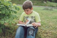 A young boy sits on grass outdoors, engrossed in reading a book.