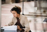 Serious young learner writing with pen on paper sheet of document for homework task in wireless headphones